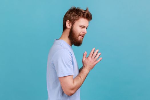 Side view of cunning bearded man schemes something keeps hands together and makes plans, sly expression looking at camera thoughtfully. Indoor studio shot isolated on blue background.