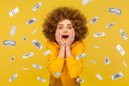 Oh my god, wow. Portrait of woman with Afro hairstyle looking at camera with big eyes, absolutely shocked of money rain falling from up. Indoor studio shot isolated on yellow background.