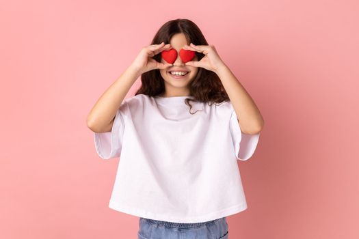 Portrait of optimistic little girl wearing white T-shirt smiling to camera, covering eyes with small red hearts, expressing love and romantic feelings. Indoor studio shot isolated on pink background.