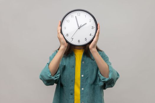 Portrait of unknown woman holding wall clock hiding her face, time management, schedule and meeting appointment, wearing casual style jacket. Indoor studio shot isolated on gray background.