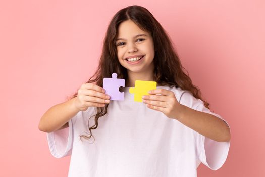 Portrait of satisfied happy smiling little girl wearing white T-shirt holding yellow and purple puzzle pieces, solving task, looking at camera. Indoor studio shot isolated on pink background.
