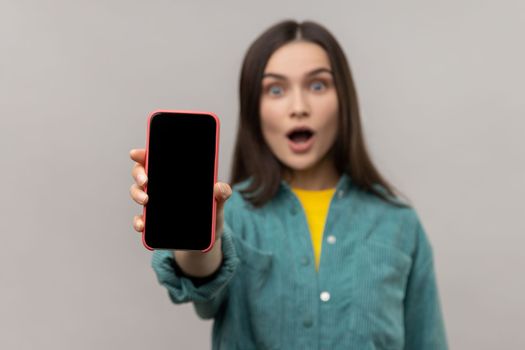 Astonished amazed woman showing cell phone with empty display with copy space for advertisement, wearing casual style jacket. Indoor studio shot isolated on gray background.