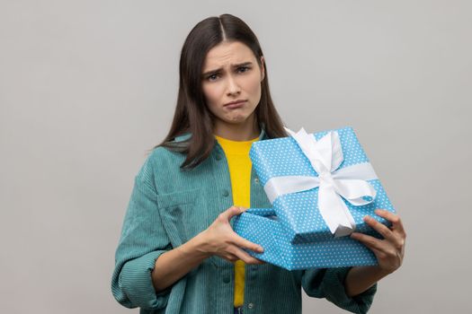 Sad upset woman with dark hair unboxing blue gift box, looking at camera with sorrow facial expression, birthday present, wearing casual style jacket. Indoor studio shot isolated on gray background.