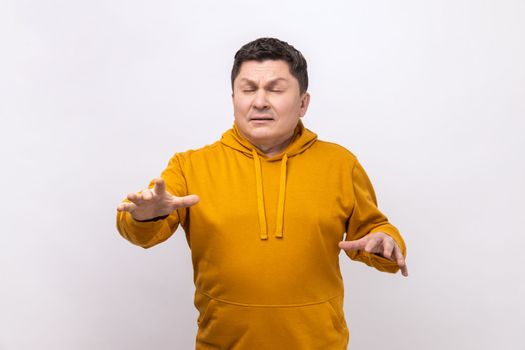 Blind unhappy man standing with closed eyes and touching air to find lost way, emulating walking in darkness, wearing urban style hoodie. Indoor studio shot isolated on white background.
