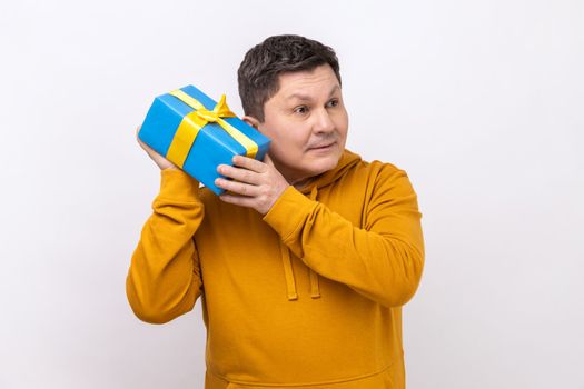 Portrait of curious middle aged man holding and shaking wrapped present box, being interested what inside, wearing urban style hoodie. Indoor studio shot isolated on white background.