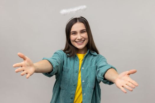 Come into my hands. Attractive angelic woman with halo over head stretching arms to camera with kind friendly smile, going to embrace. Indoor studio shot isolated on gray background.