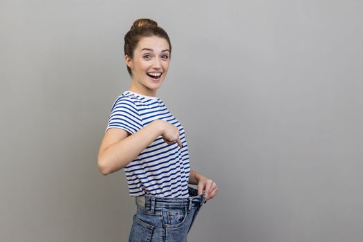 Attractive woman wearing striped T-shirt have willpower to loose weight, pointing at slim waist and pointing finger on big trousers, diet conception. Indoor studio shot isolated on gray background.