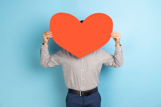 Anonymous date. Portrait of man hiding his face behind big read paper heart, holding symbol of love, care, generosity, give hope, wearing striped shirt. Indoor studio shot isolated on blue background.