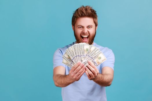 Portrait of extremely happy bearded man holding lot of money and screaming to camera, showing dollar bills in hand, enjoying financial success. Indoor studio shot isolated on blue background.