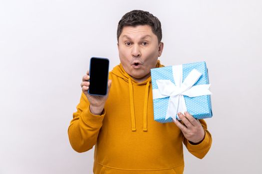Amazed man holding blue present box and smatrphone with blank screen for promotion, looking at camera with open mouth, wearing urban style hoodie. Indoor studio shot isolated on white background.
