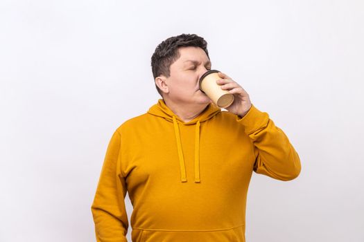 Portrait of handsome middle aged man standing and drinking take away coffee in paper cup, freshness lifestyle, wearing urban style hoodie. Indoor studio shot isolated on white background.