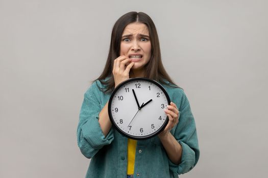 Portrait of impatient nervous woman with dark hair biting her nails and holding big wall clock, deadline, wearing casual style jacket. Indoor studio shot isolated on gray background.