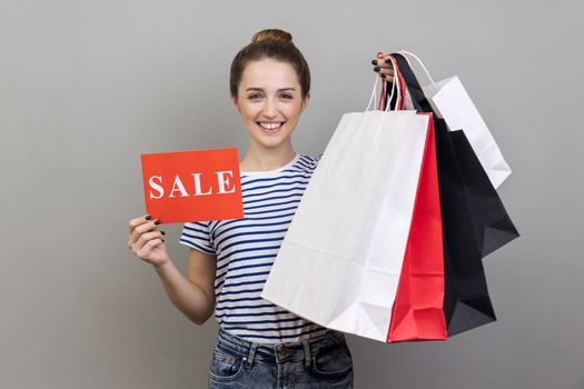 Portrait of smiling satisfied woman wearing striped T-shirt with purchases holding paper shopping bags and sale card, big holidays discounts. Indoor studio shot isolated on gray background.