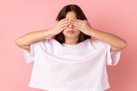 Portrait of sad upset little girl wearing white T-shirt standing covering eyes with hand, does not want see at something shameful. Indoor studio shot isolated on pink background.