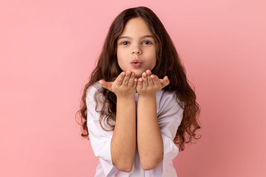 Portrait of romantic lovely little girl wearing white T-shirt sending air kisses to her friend, looking at camera, falling in love. Indoor studio shot isolated on pink background.