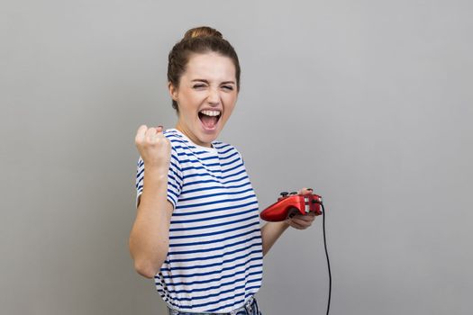 Portrait of woman wearing striped T-shirt holding gamepad, winning video games competition, clenched fist with happy expression, rejoices victory. Indoor studio shot isolated on gray background.