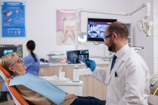 Dentist assistant giving doctor radiography of senior woman waiting. Medical teeth care taker holding patient radiography sitting on chair during consultation.