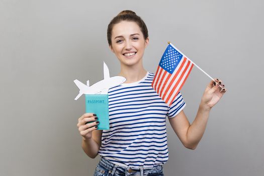 Portrait of smiling woman wearing striped T-shirt standing with American flag and passport with paper plane, travel abroad. Indoor studio shot isolated on gray background.