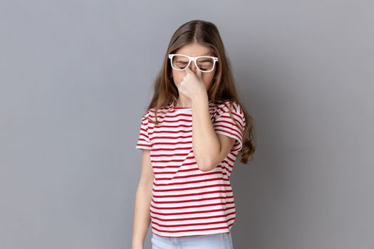 Portrait of sick tired little girl wearing striped T-shirt standing ribbing her eyes, takes off glasses, felling eyes hurt and headache. Indoor studio shot isolated on gray background.