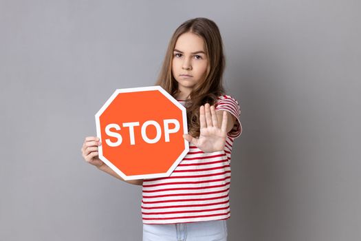 Little girl in T-shirt covering face with octagonal Stop symbol, showing red traffic sign and ban gesture with palm, warning of forbidden way. Indoor studio shot isolated on gray background.