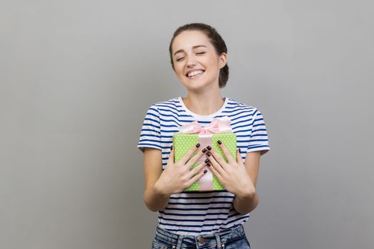 Portrait of happy smiling pleased woman wearing striped T-shirt embracing green wrapped present box, being satisfied of such a great gift. Indoor studio shot isolated on gray background.