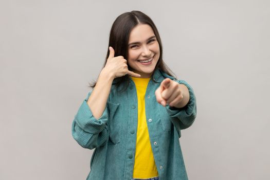 Hey you, call me back, let's contact by phone. Portrait of playful happy adult woman making telephone gesture and pointing to camera. Indoor studio shot isolated on gray background.