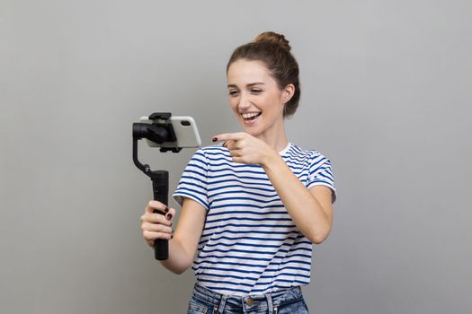 Portrait of attractive woman wearing striped T-shirt pointing to cell phone camera with smile, streaming, talking with followers, uses steadicam. Indoor studio shot isolated on gray background.