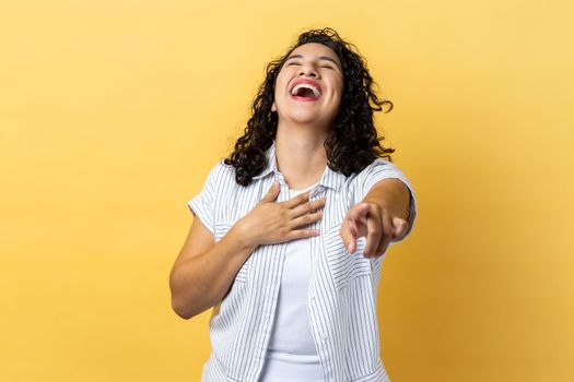 Portrait of happy woman with dark wavy hair laughing out loud and pointing finger to camera, hearing funny joke, holding belly. Indoor studio shot isolated on yellow background.