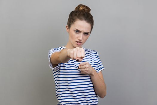 Portrait of aggressive woman wearing striped T-shirt standing holding clenched fists up ready to boxing, fighting spirit, boxing. Indoor studio shot isolated on gray background.