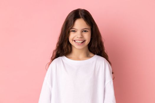 Portrait of happy satisfied little girl wearing white T-shirt looking at camera with toothy smile, expressing positive expression. Indoor studio shot isolated on pink background.