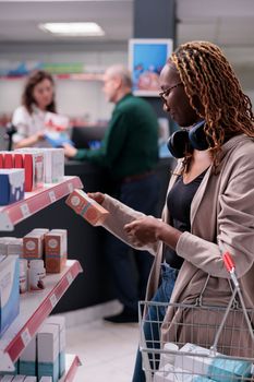 Customer looking at shelves full with pharmaceutical products, buying supplements after reading medication leaflet. Clients holding baskets putting pills package in it. Health care service