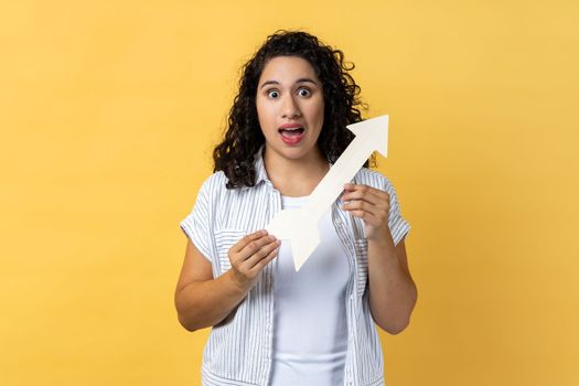 Portrait of amazed excited woman with dark wavy hair standing holding white arrow pointing aside, looking at camera with open mouth. Indoor studio shot isolated on yellow background.