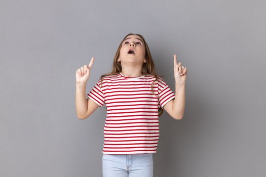 Wow look there, advertising area. Little girl looking astonished and pointing up copy space, showing empty place above head for promotion. Indoor studio shot isolated on gray background.
