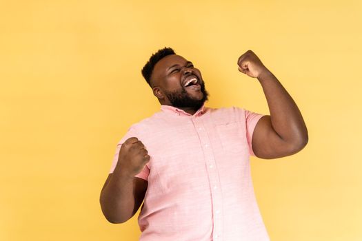 Portrait of extremely happy man wearing pink shirt celebrating his success, standing with clenched fists, screaming happily. Indoor studio shot isolated on yellow background.