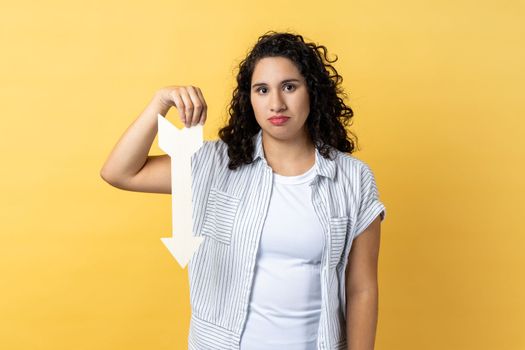Portrait of upset dissatisfied woman with dark wavy hair showing white arrow pointing down, expressing sad emotions, downgrade concept. Indoor studio shot isolated on yellow background.