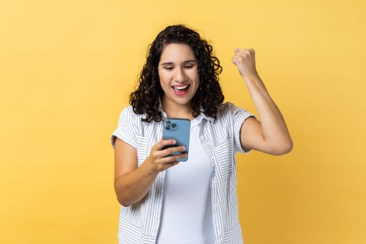 Portrait of extremely happy woman with dark wavy hair holding smart phone and clenched fist, showing yes gesture, celebrating her winning. Indoor studio shot isolated on yellow background.