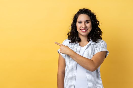 Portrait of smiling satisfied beautiful woman with dark wavy hair standing pointing aside with finger, showing copy space for advertisement. Indoor studio shot isolated on yellow background.