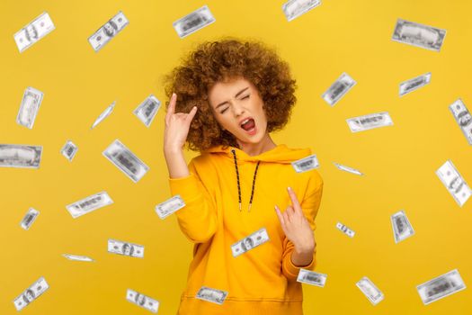 Portrait of woman with Afro hairstyle wearing casual style hoodie showing rock and roll, rejoicing money rain falling from up, winning lottery. Indoor studio shot isolated on yellow background.