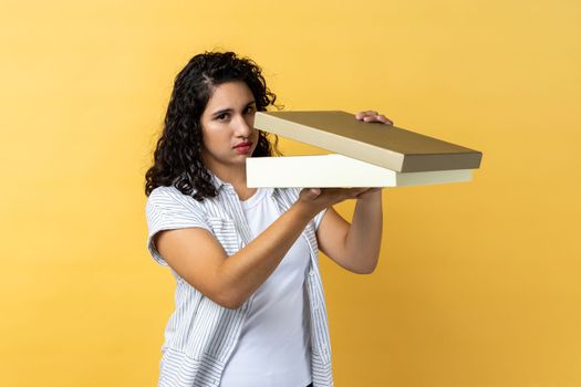 Portrait of sad upset woman with dark wavy hair opens wrapped present box, expressing disappointment, looking at camera, expressing sorrow. Indoor studio shot isolated on yellow background.