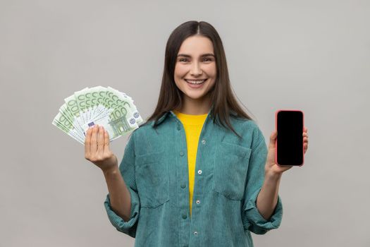 Happy positive rich woman holding euro banknotes and phone with blank screen for advertisement or promotion, wearing casual style jacket. Indoor studio shot isolated on gray background.