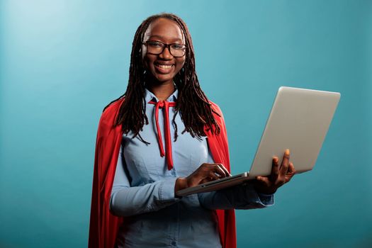 Positive and happy superhero woman wearing mighty hero red cape surfing internet on modern laptop while looking at camera. Brave justice defender with superpowers browsing web on handheld computer