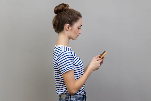 Side view portrait of serious woman wearing striped T-shirt holding smartphone, typing message or browsing network, communicating using cell phone. Indoor studio shot isolated on gray background.