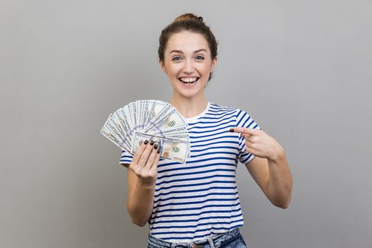 Portrait of amazed positive woman wearing striped T-shirt pointing at fan of dollar banknotes, showing big money and looking at camera. Indoor studio shot isolated on gray background.