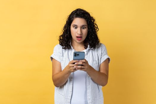 Portrait of shocked astonished woman with dark wavy hair using mobile phone and reading shocking news in social news, looking at screen with big eyes. Indoor studio shot isolated on yellow background.