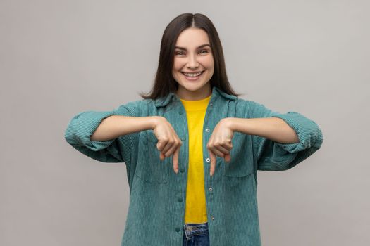 Happy woman with dark hair pointing fingers down showing space for your advertisement, looking at camera with toothy smile, wearing casual style jacket. Indoor studio shot isolated on gray background.