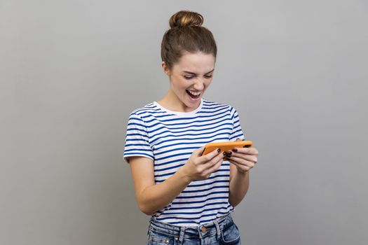 Portrait of happy woman wearing striped T-shirt using mobile phone with attentive focused expression, playing video game on cellphone. Indoor studio shot isolated on gray background.