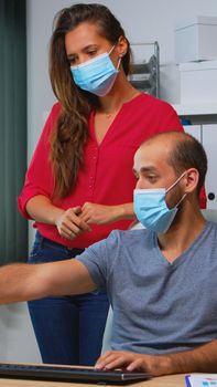 Coworkers with protection face masks working together in workplace during pandemic. Team in new normal office workspace in personal corporate company typing on computer keyboard looking at desktop