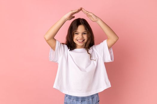 Portrait of little girl wearing white T-shirt with charming smile holding hands in gesture of house roof over head and smiling, feeling safe. Indoor studio shot isolated on pink background.