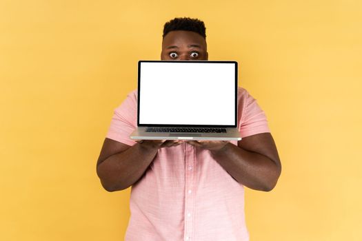 Man wearing pink shirt covering half of face with laptop with blank white screen and looking at camera with excited facial expression and big eyes. Indoor studio shot isolated on yellow background.