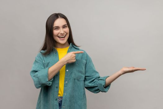 Portrait of happy woman presenting copy space on her palm, showing empty place for commercial text or goods, wearing casual style jacket. Indoor studio shot isolated on gray background.
